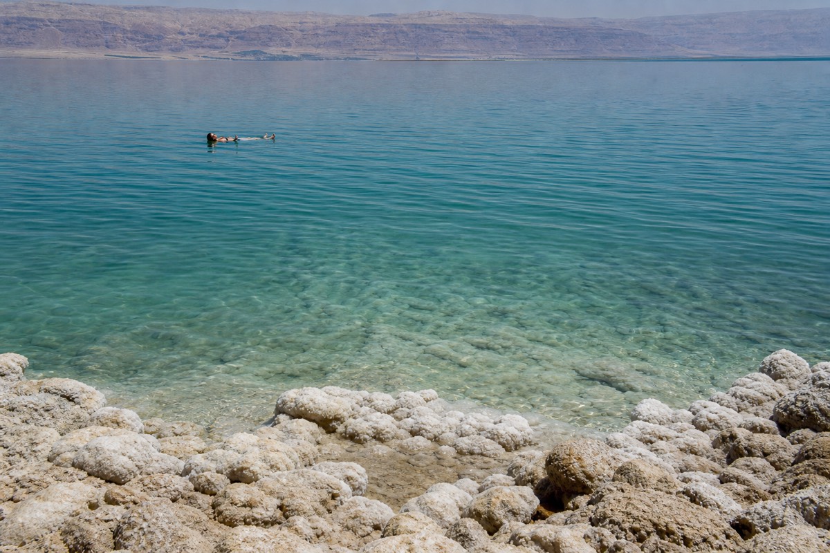 A traveler floating effortlessly on the surface of the Dead Sea with salt-crusted rocks visible along the mineral-rich shoreline